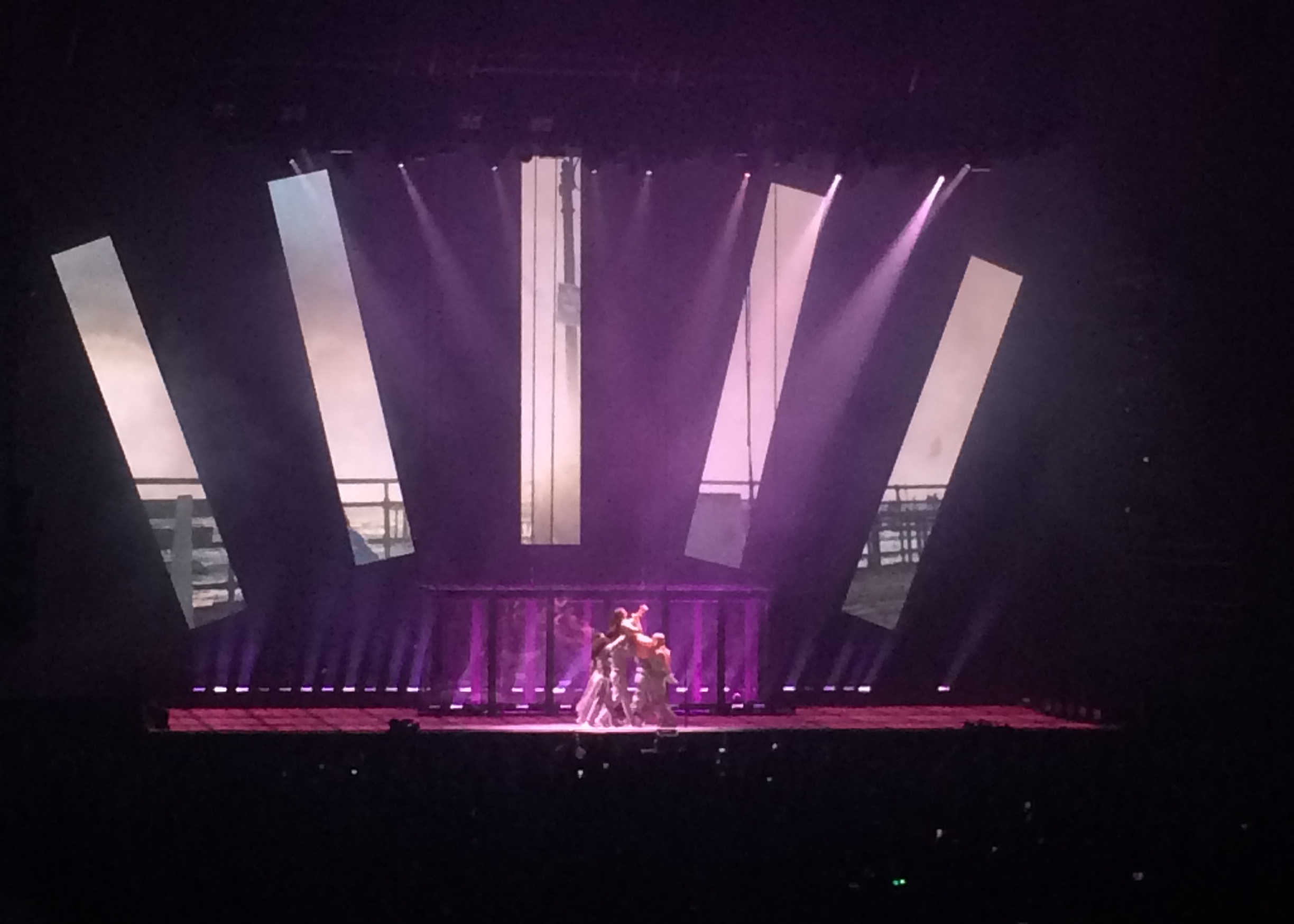 Stage photo of Lorde being lifted and carried by a small group of dancers underneath a large projected display of five lighted panels fanned out. The panels are displaying a broken up image of a ship at sea.