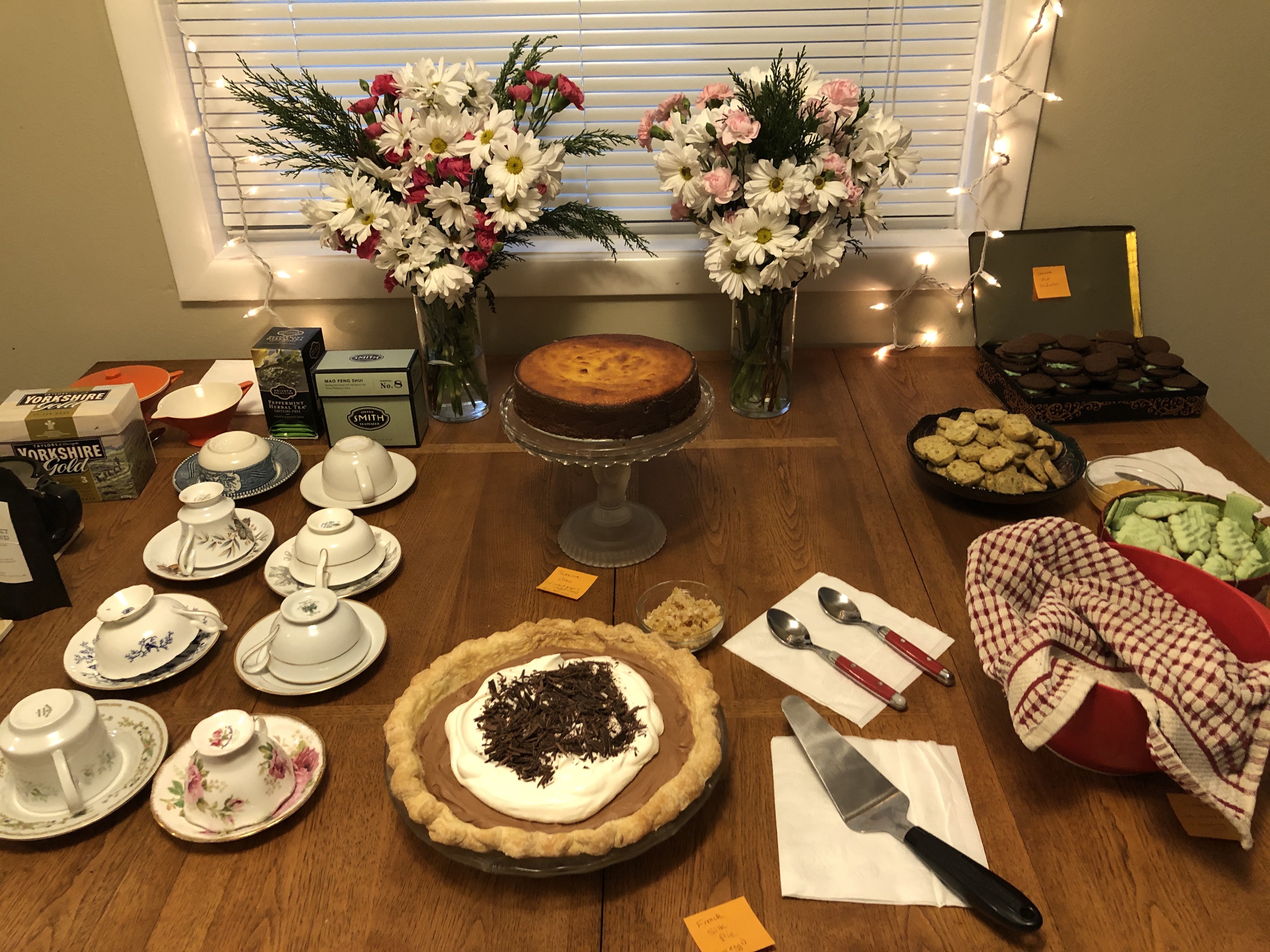 A table filled with desserts. There's an orange cake on a glass cake stand, a chocolate pie, three tins of Christmas cookies, two vases of fresh flowers, and a line-up of teacups and tea to the side.