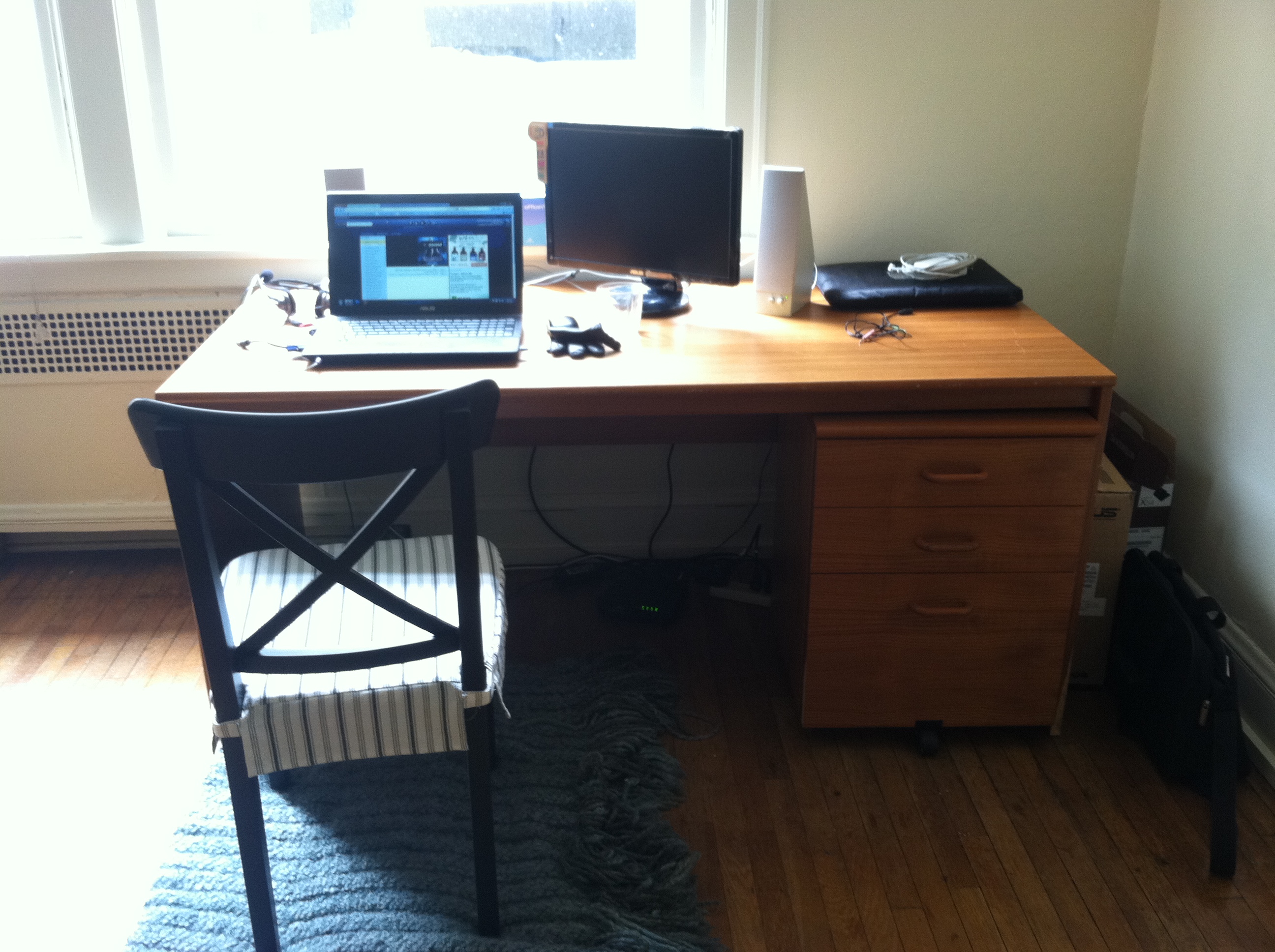 Large wooden desk in the corner of a bare studio apartment. It has a laptop and small monitor on it. The mouse wristrest is an old rolled-up glove. The chair is a black kitchen chair from Ikea. Through the window you can make out the window frame of the next door apartment I faced into.