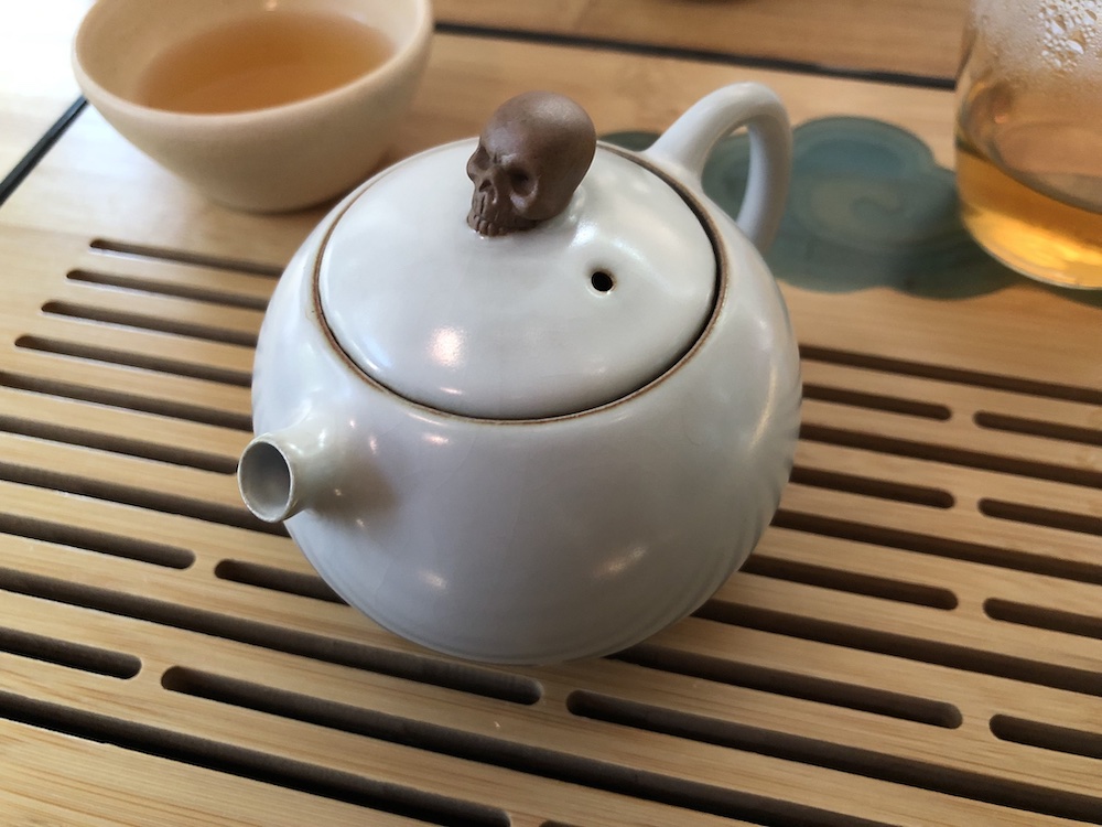 A small xi shi style teapot. It is the size and shape of an orange, with a white ruyao glaze that will crackle over time. The teapot knob is a skull in unglazed reddish clay.