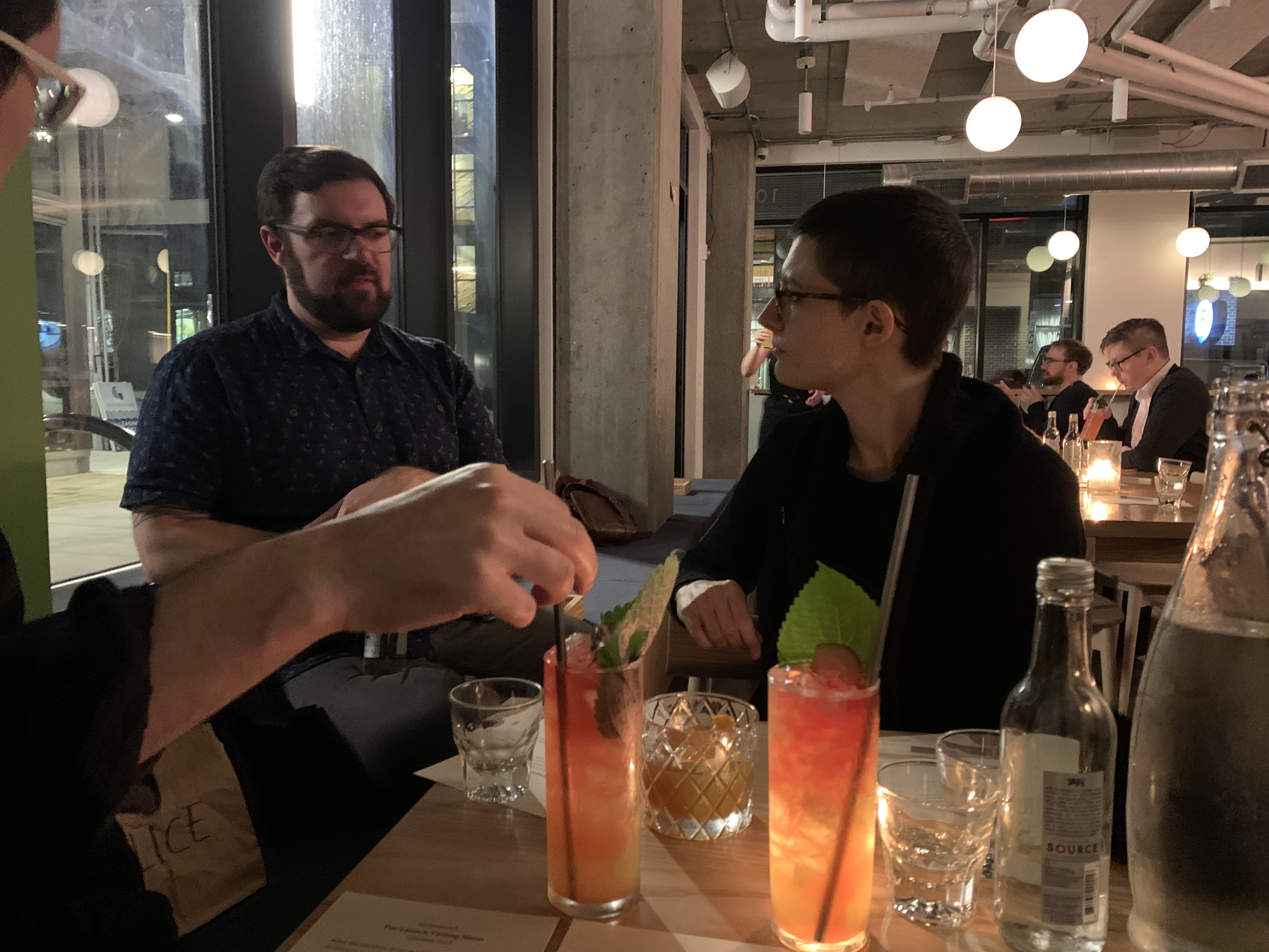 Photo of me from across a small restaurant table, indoors. I am in a black sweater and turned to listen to a white man with short hair and glasses.