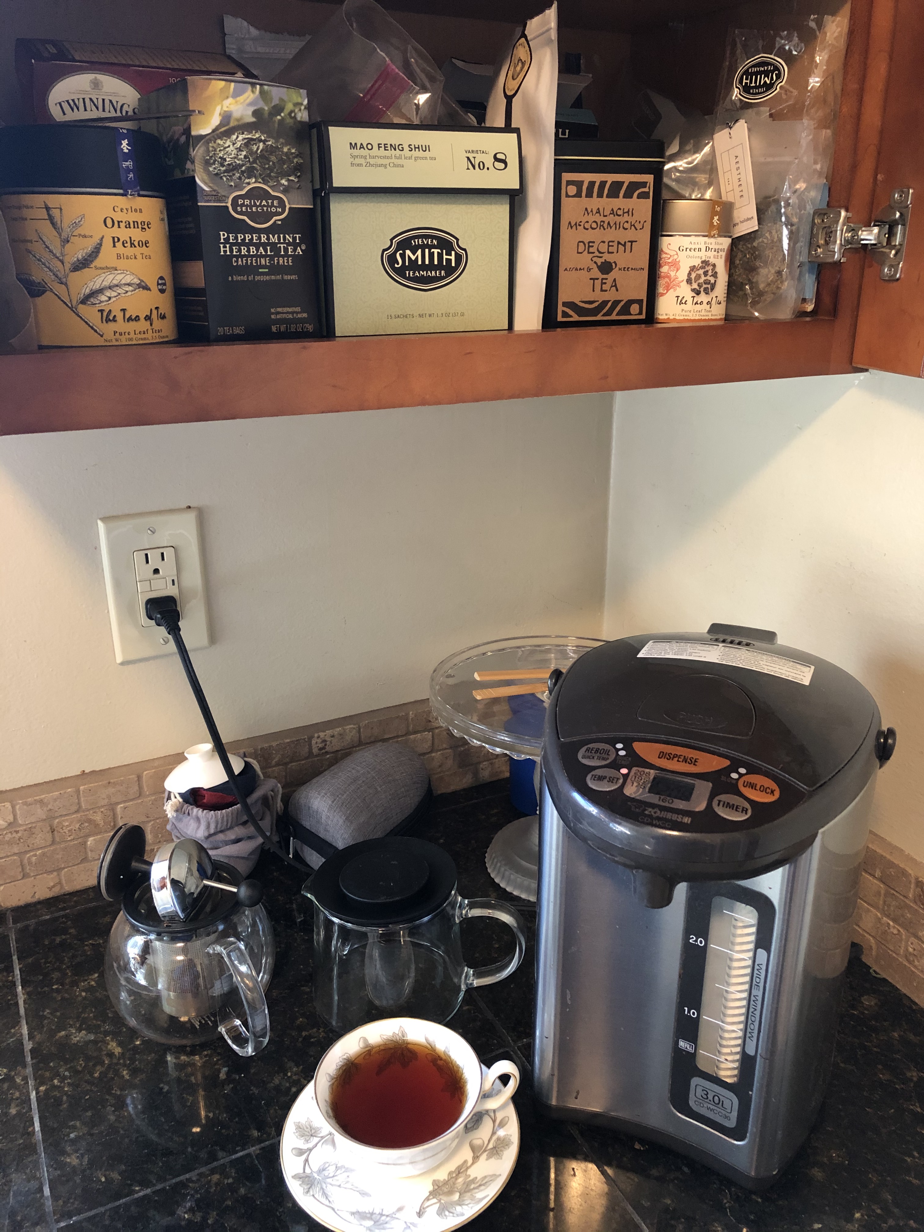 My home tea station. Corner of a kitchen counter with a water boiler, two glass teapots, and a porcelin teacup with tea in it. Above the counter is an open cabinet showing a variety of looseleaf and bagged teas.